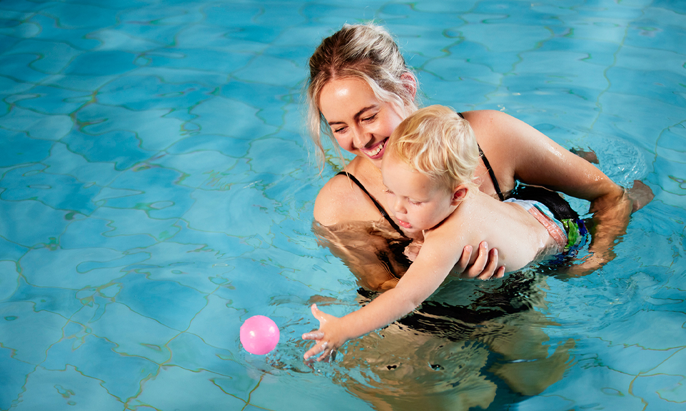 Smiling mum holding baby playing with ball in pool